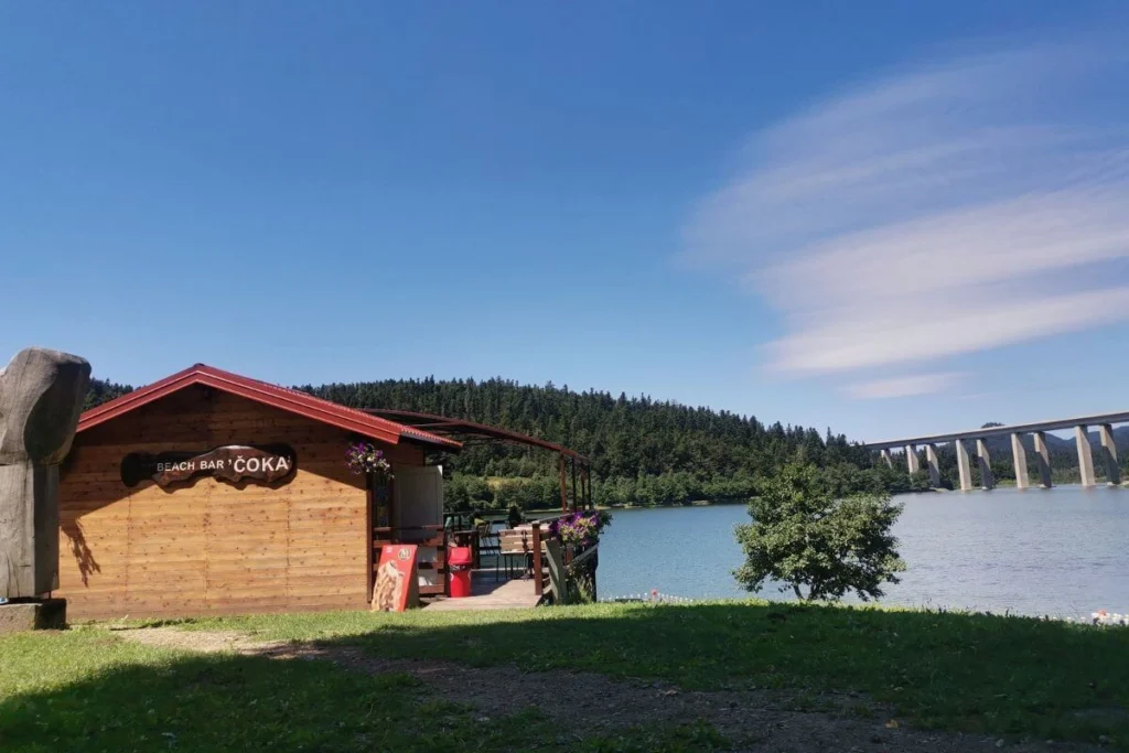 Beach bar Čoka on the shore of Lake Bajer in Fužine, with the tall A1 highway bridge in the distance and forested mountains.