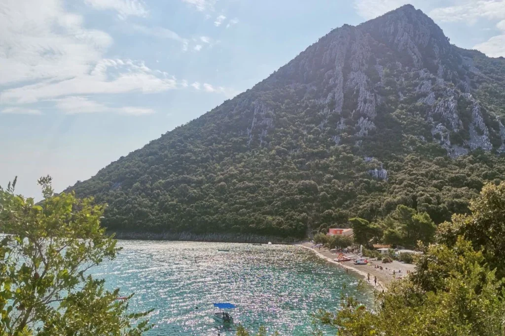Wide view of Divna Bay on the Pelješac Peninsula featuring a pebble beach, turquoise water, and a dramatic green mountain rising directly from the shore.
