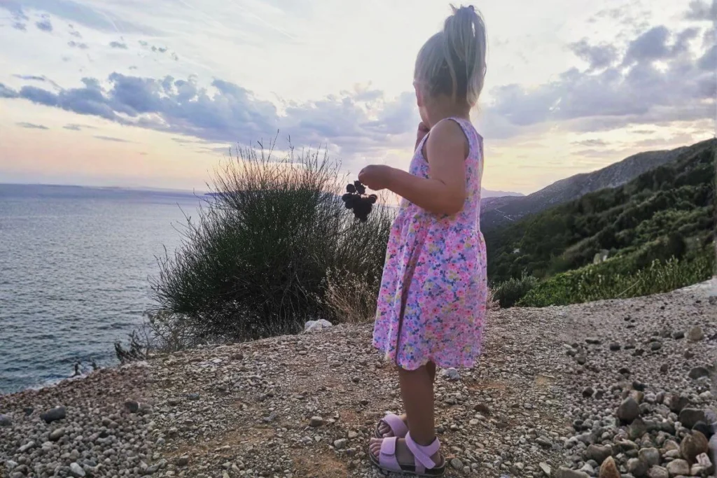 A young girl in a floral dress holding a bunch of purple grapes, standing on a gravel path overlooking the Adriatic Sea and the steep vineyards of the Pelješac Peninsula.