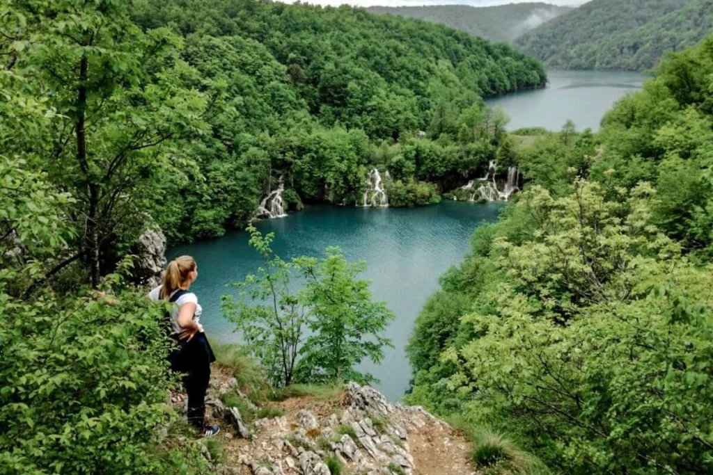 Woman with a baby carrier looking out over the turquoise waters and waterfalls of Plitvice Lakes National Park, showcasing the steep, wooded terrain.