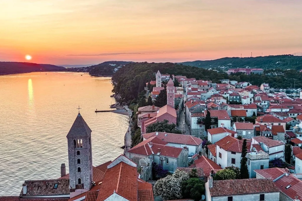 Aerial sunset view of the historical old town of Rab on Rab Island, Croatia, featuring red-roofed stone houses and iconic church bell towers along the Adriatic coast.