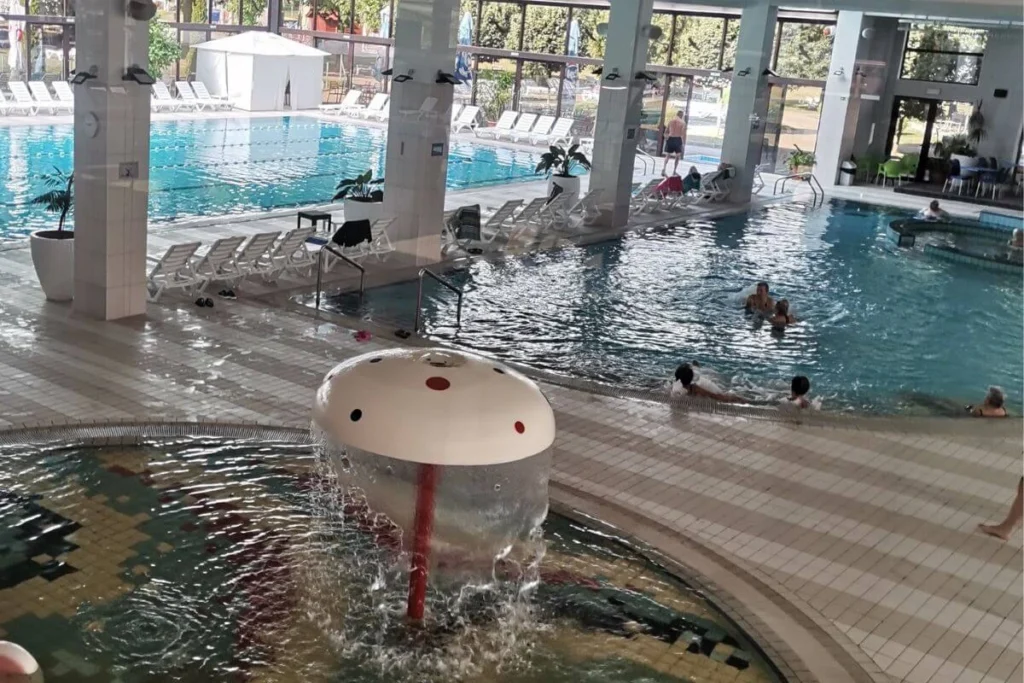 Large indoor thermal pool complex at Terme Jezerčica with a shallow children's pool featuring a mushroom water fountain and lounge chairs for parents.