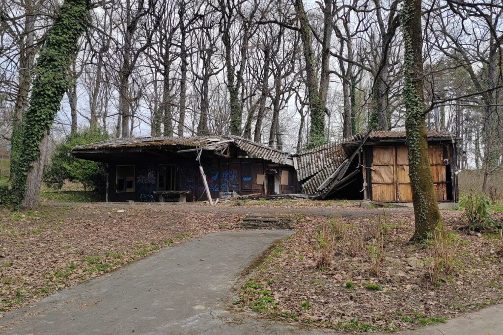 A weathered wooden pavilion with a steep triangular roof, showing signs of decay and aging, tucked away in the forest of Grad Mladih.