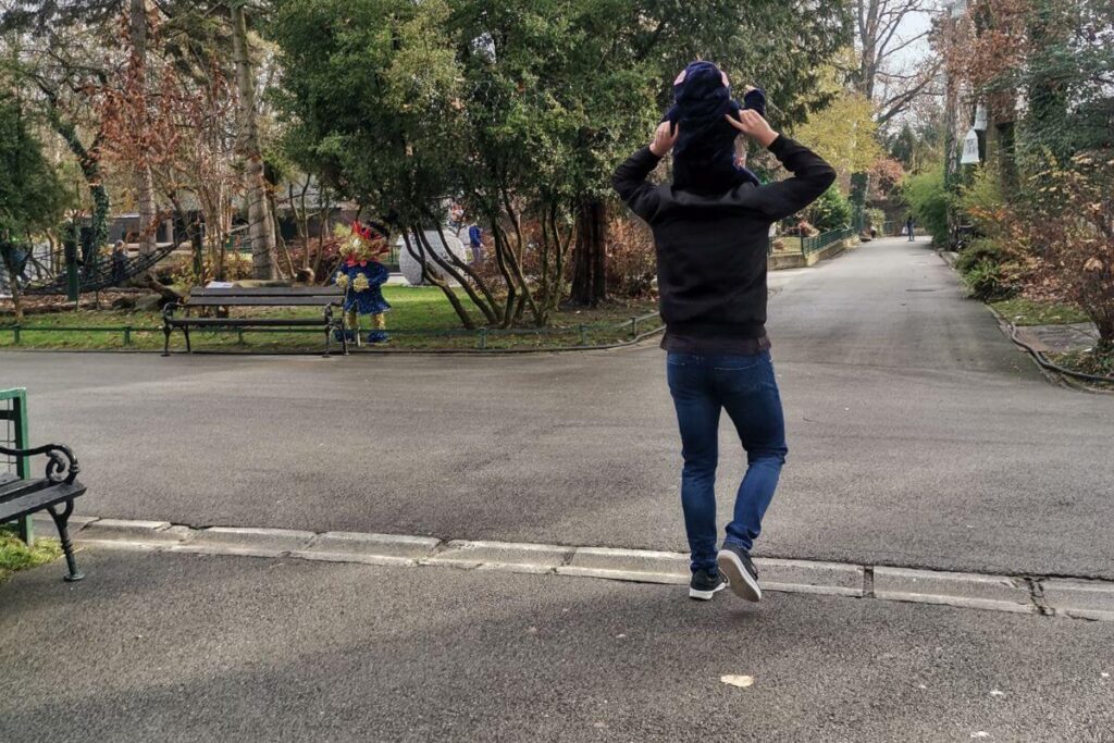 Father walking on a wide paved path at Zagreb Zoo with a toddler on his shoulders.