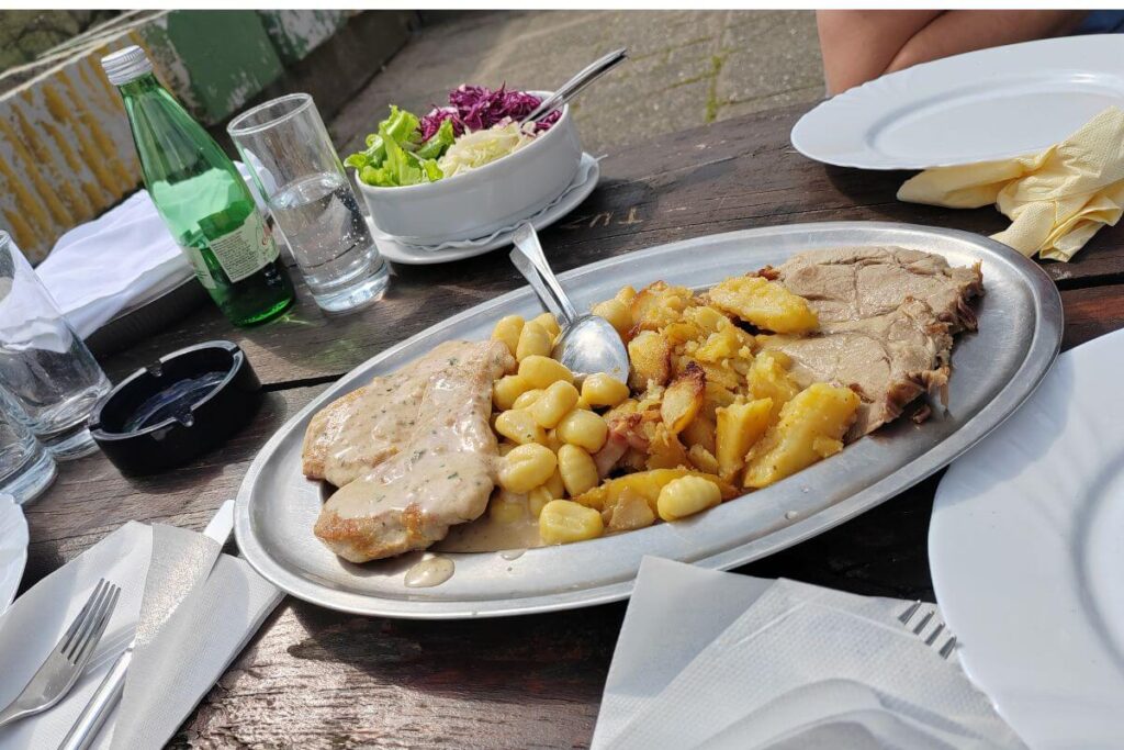 A large metal platter filled with roasted meat, potatoes, and gnocchi in sauce, served on a wooden table at the Grad Mladih restaurant.