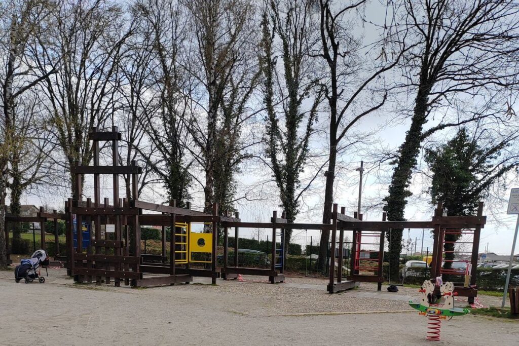 A child climbing on a large, multi-level dark wooden adventure playground structure surrounded by a pine forest at Grad Mladih in Zagreb.