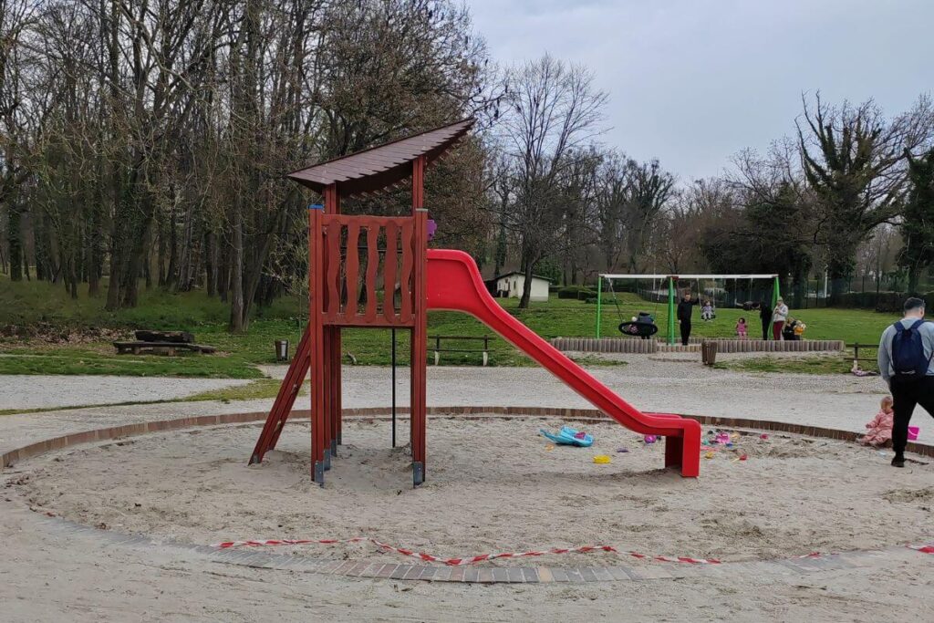  A red plastic slide attached to a wooden play tower, situated in the center of a large, circular sandpit at the Grad Mladih playground in Zagreb. 