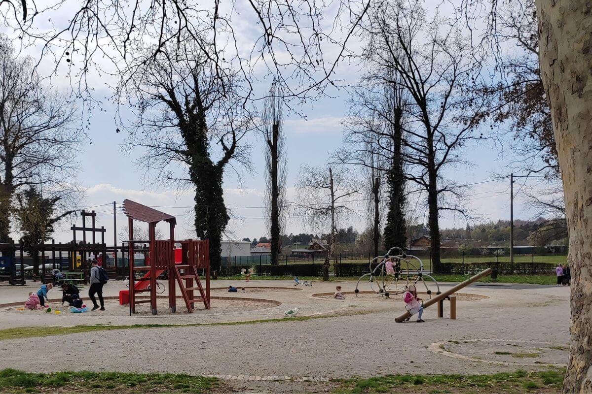 A wide view of the main playground at Grad Mladih Zagreb, showing children playing on large wooden climbing frames, a sandpit, and a see-saw under tall trees.