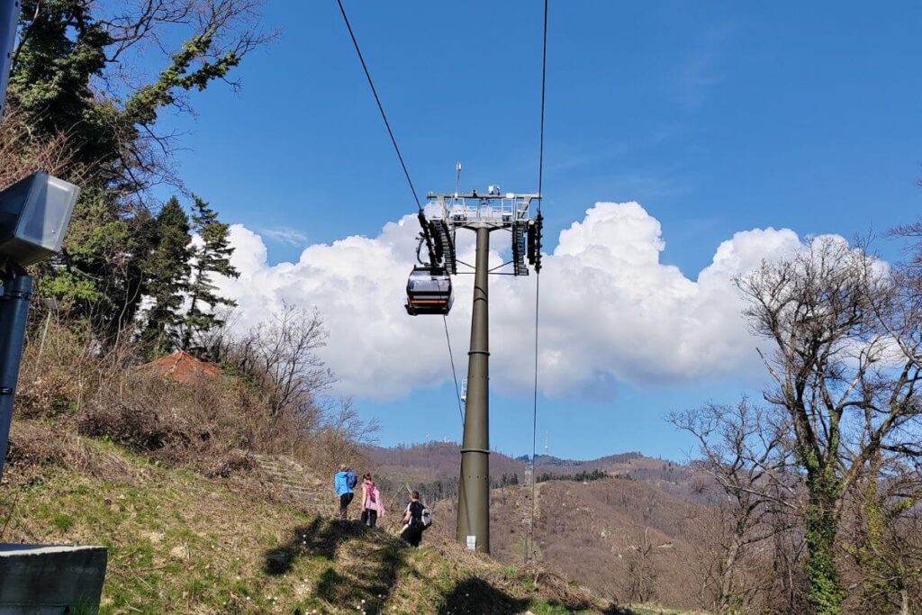 A sleek, modern black gondola of the Sljeme cable car (Žičara) ascending a tall green support pillar over a lush green forest toward the mountain summit under a blue sky