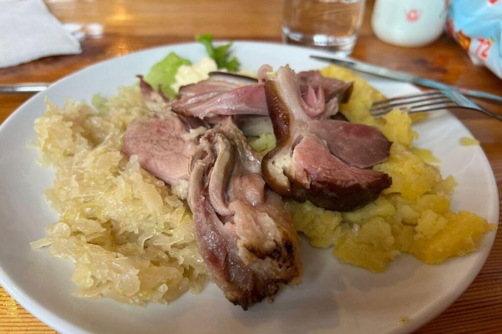 A white plate filled with a traditional Croatian meal consisting of a large piece of roasted meat, a serving of sauerkraut, and creamy mashed potatoes, served in a rustic mountain hut setting.