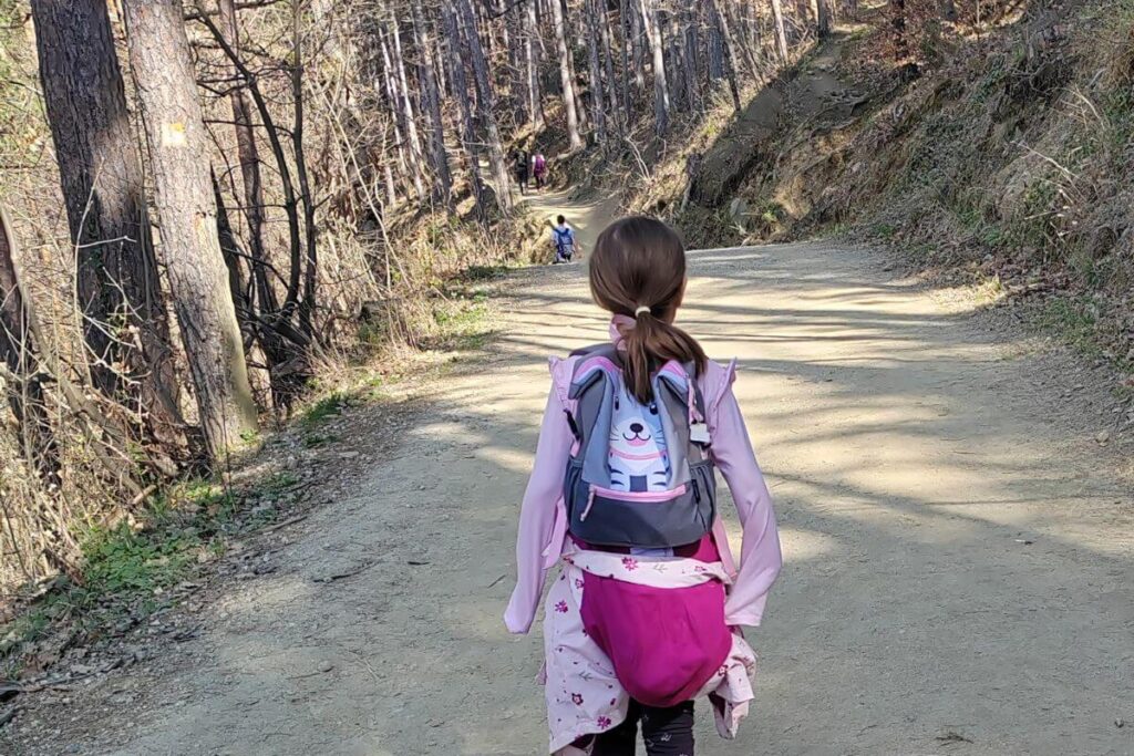 ear view of a young girl with a ponytail wearing a pink and grey bunny-themed backpack walking down a wide, sun-dappled forest trail on Mount Sljeme.