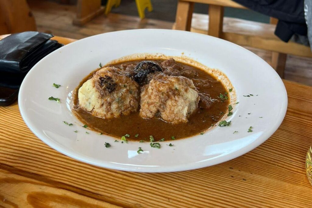A close-up of a traditional Croatian mountain meal featuring a white bowl of thick bean stew with two large dumplings and sliced sausages, served on a rustic wooden table.