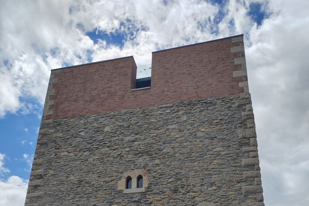 A close-up view looking up at a tall, historic stone fortress tower with a newly restored red brick top section against a blue sky.