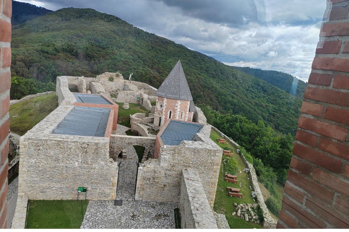 An elevated viewpoint looking down into the stone courtyard, chapel, and protective walls of Medvedgrad fortress surrounded by green hills.