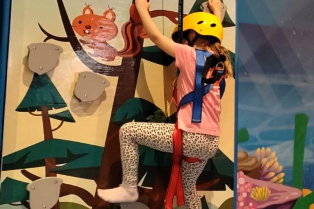 A young girl wearing a yellow safety helmet and harness climbs an indoor rock climbing wall at Family Hotel Lišanj.