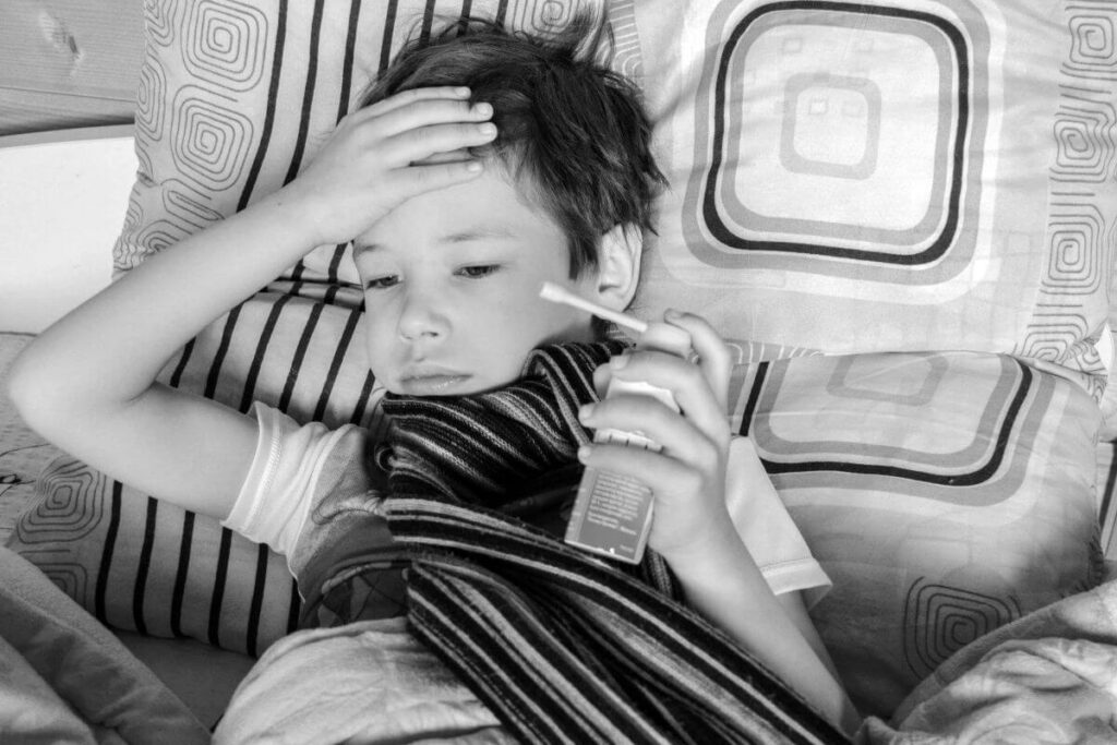 A young boy lying in bed with a striped scarf, holding a medicine spray and touching his forehead due to a fever.