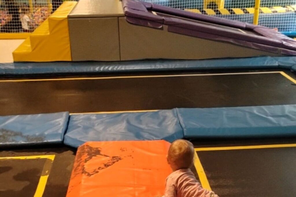 A baby crawling on soft safety mats toward an indoor trampoline and active play structure.