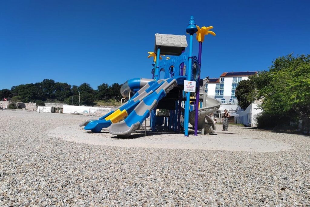 A large blue and yellow plastic playground structure with slides on a wide pebble beach under a clear blue sky.