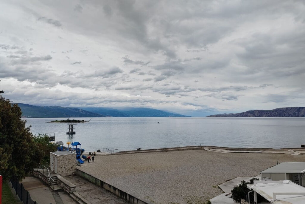 Panoramic sea view from a balcony overlooking a pebble beach and the small island of San Marino under a cloudy sky.