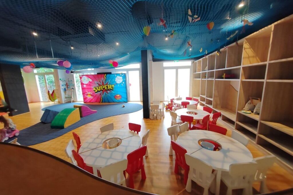 A spacious indoor kids club room featuring small round white tables, colorful red chairs, and creative play areas.