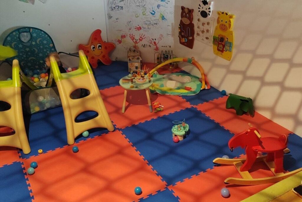 A soft-play indoor nursery corner with puzzle floor mats, plastic slides, toys, and a wooden rocking horse.