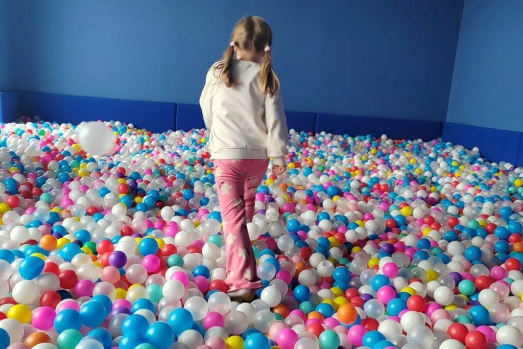 A young girl with pigtails from behind walking through a massive room filled with colorful plastic ball pit balls.