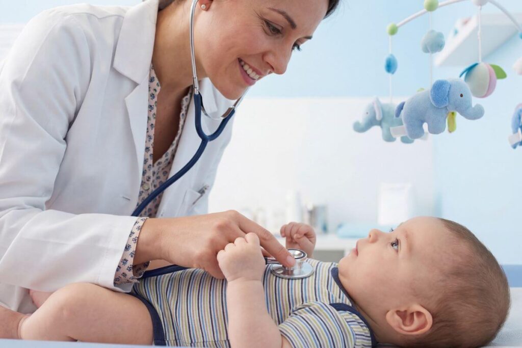 A friendly female pediatrician smiling warmly while checking a baby's heartbeat with a stethoscope in a bright clinic room.