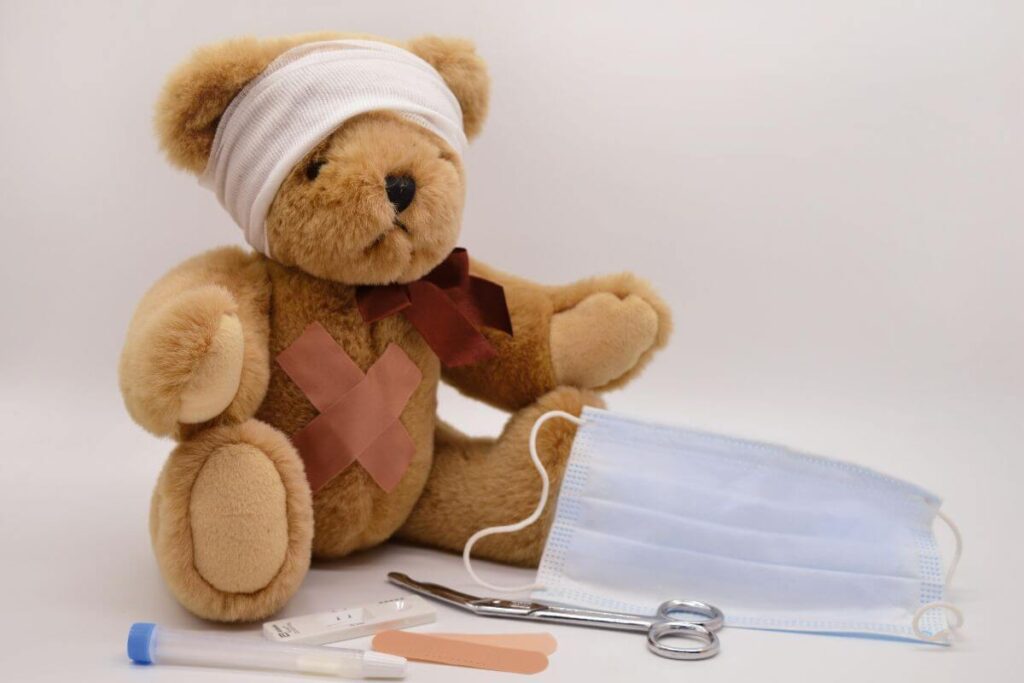 A plush teddy bear sitting with a bandage wrapped around its head, a cross-shaped band-aid on its chest, next to a medical face mask and scissors.