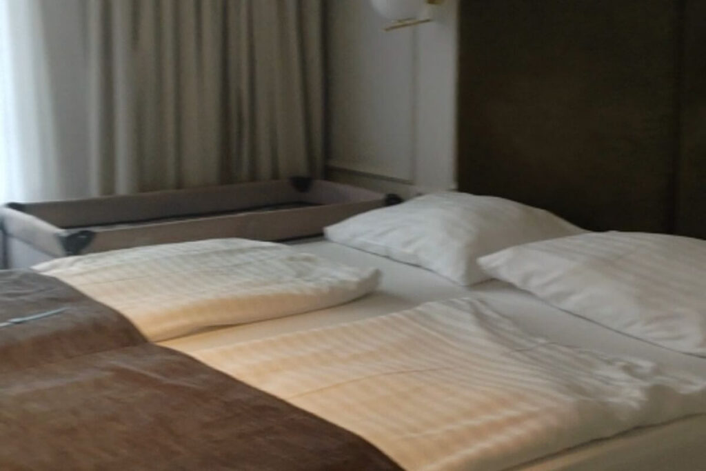 A clean double hotel bed with white linens and a travel baby cot set up in the background by the window.