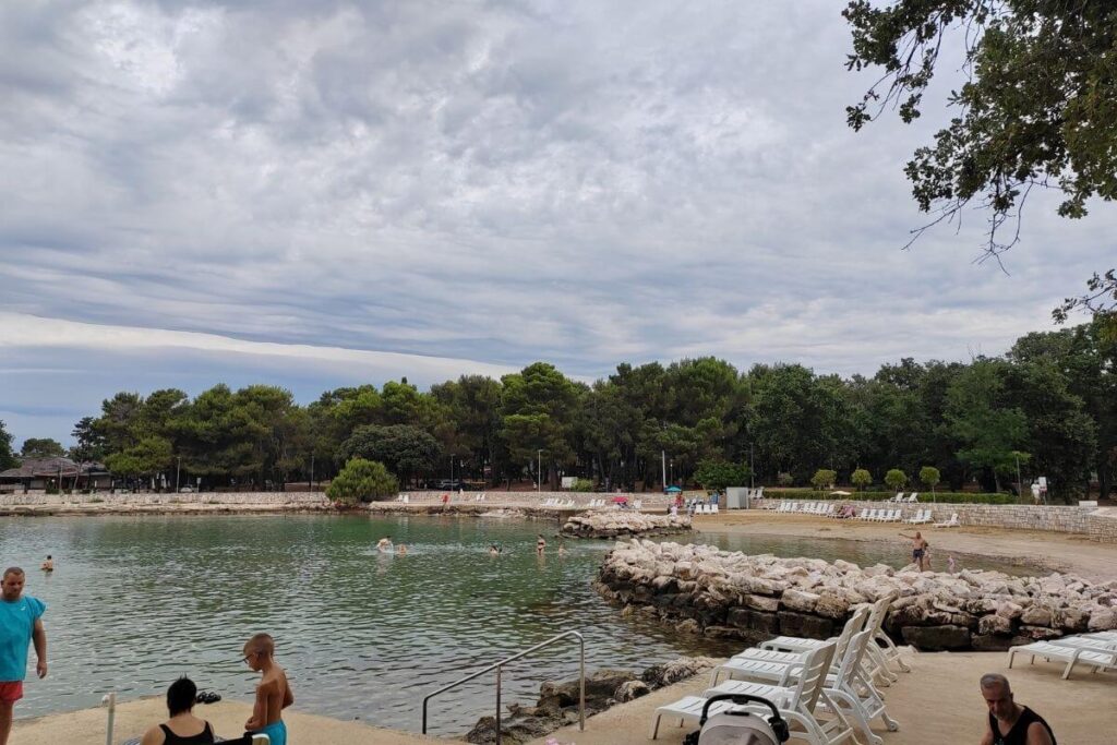 Sheltered sea cove with calm green water, rock breakwaters, paved sunbathing terraces, and a stroller parked by sunbeds.