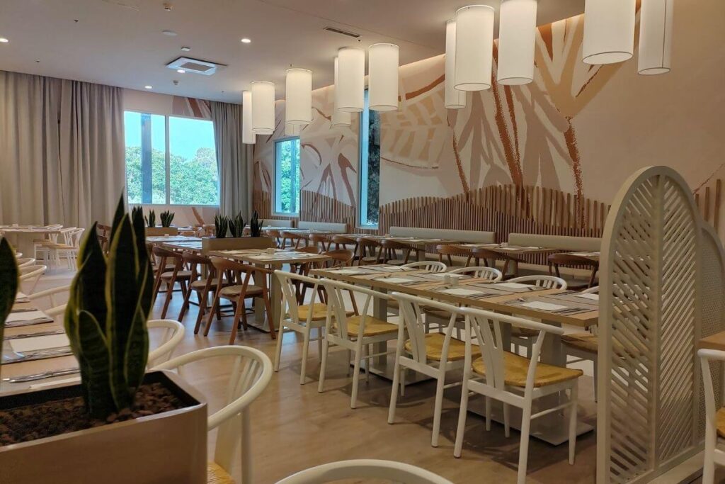 Bright modern hotel restaurant dining hall with wooden tables, white chairs, soft cylindrical lighting, and natural decor accents.