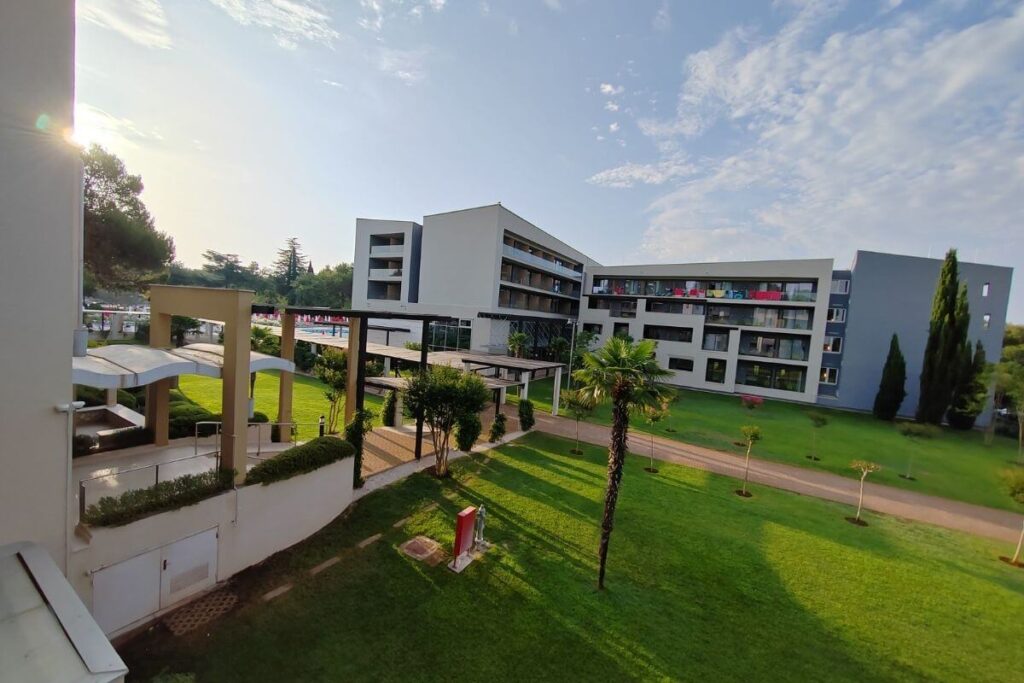 High-angle view from a hotel balcony showing lush green lawns, palm trees, paved walking paths, and modern hotel accommodation wings.