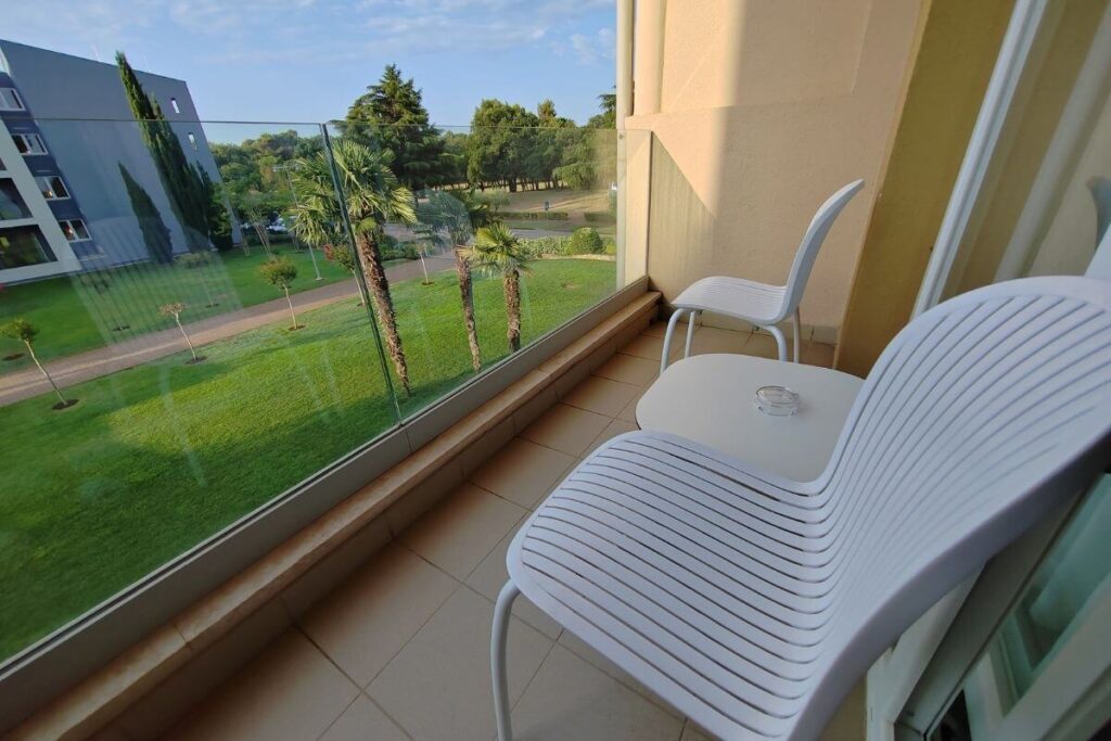Private hotel room balcony with white modern chairs, a small table, and a protective glass safety railing overlooking green trees.