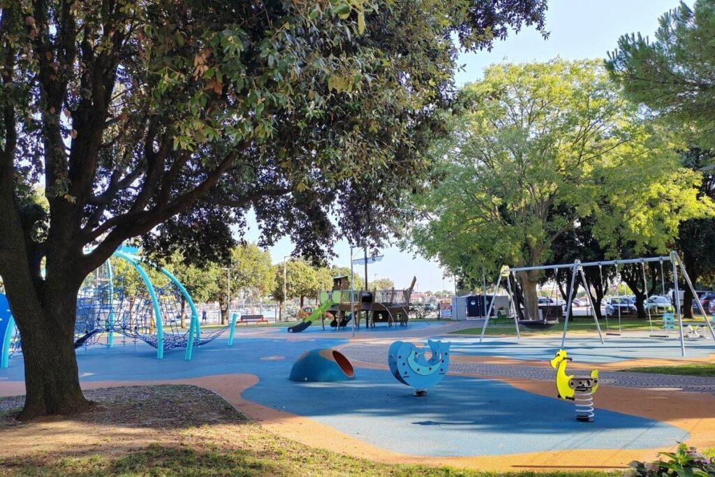Large public playground in Umag town center with swings, climbing nets, slides, and safety rubber flooring under shade trees.
