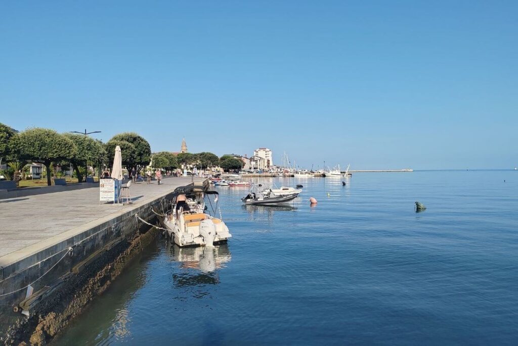 Paved coastal promenade alongside calm blue sea water with small boats moored along the stone pier in Umag town center.