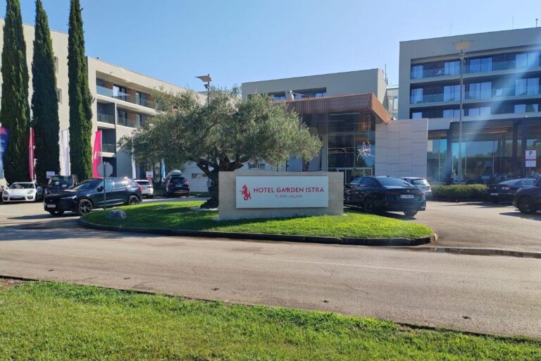 Front entrance sign for Hotel Garden Istra Plava Laguna with landscaped grass, olive tree, parked cars, and hotel facade.