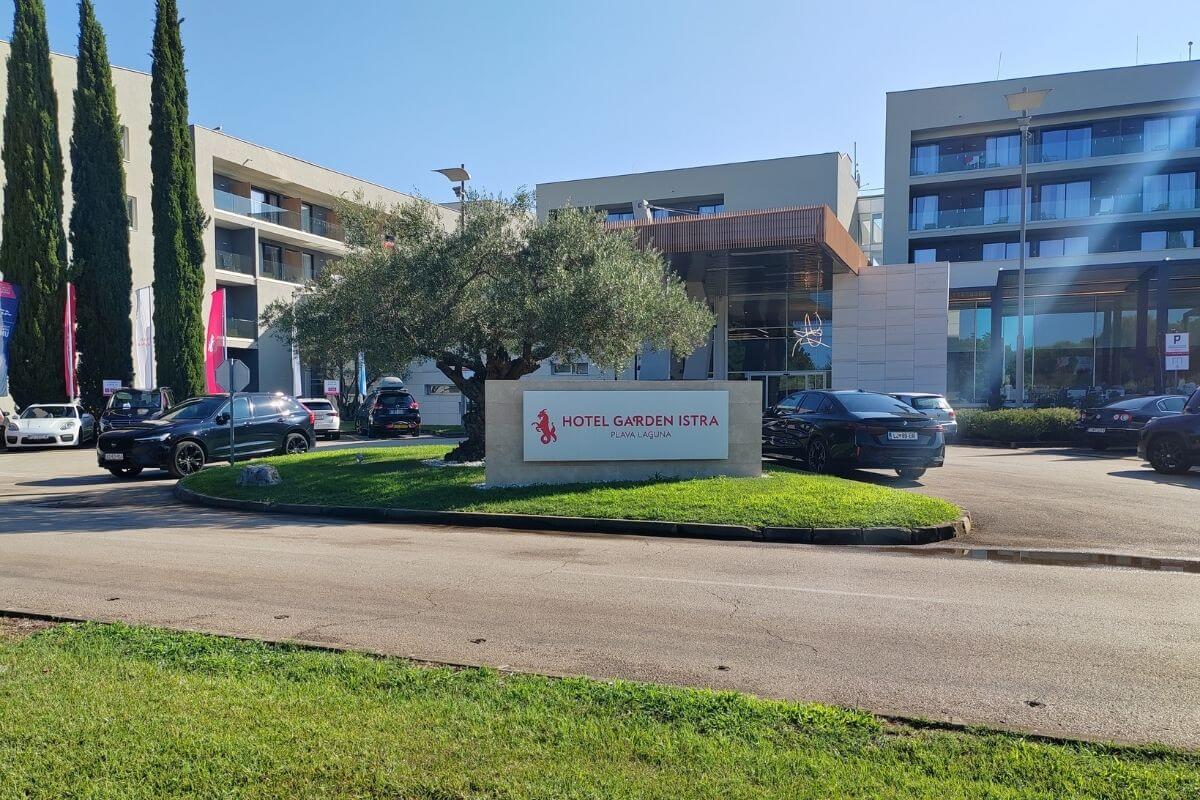Front entrance sign for Hotel Garden Istra Plava Laguna with landscaped grass, olive tree, parked cars, and hotel facade.