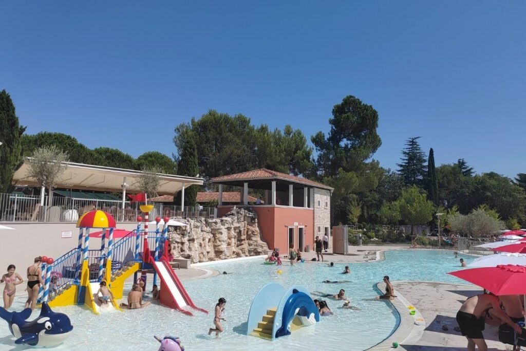 Children playing in a shallow outdoor splash pool featuring colorful water slides, play structures, and rock waterfall accents.
