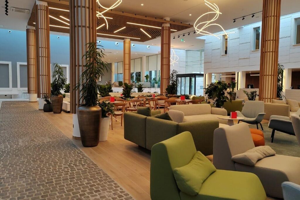 Spacious, open-concept hotel lobby lounge featuring modern green armchairs, wooden columns, potted plants, and modern light fixtures.
