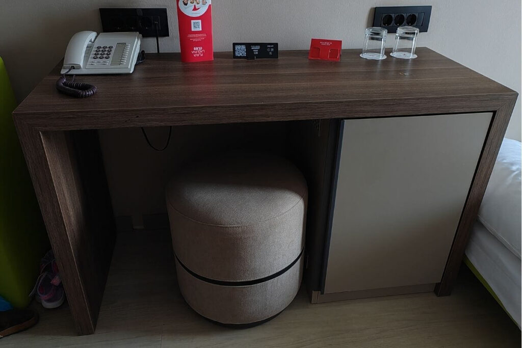 Dark wood room desk featuring a mini-fridge cabinet, ottoman stool, telephone, and wall power outlets.