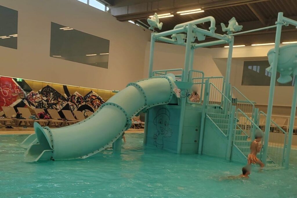 Indoor children's shallow pool with a light blue tube slide and interactive water spray features.