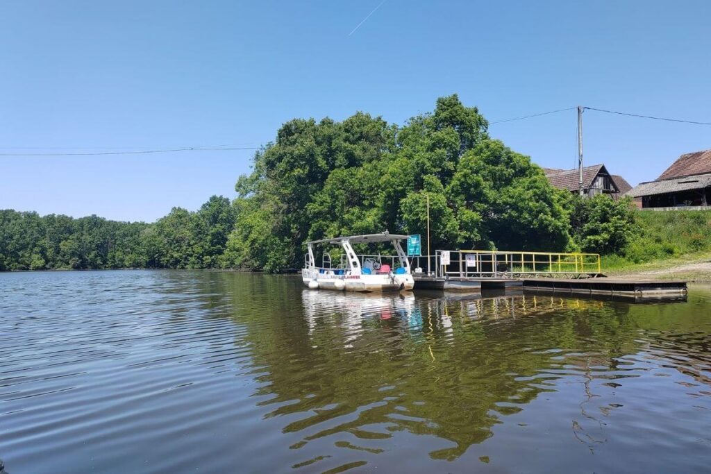 Solar-powered boat docked at a wooden pier on the river in Lonjsko Polje with traditional houses in the background   